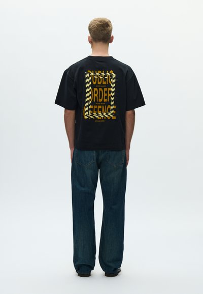 Μαύρο υπερμεγέθες μπλουζάκι με κίτρινο γραφικό κείμενο και ριγωτή μπορντούρα στην πλάτη, συνδυασμένο με φαρδιά μπλε τζιν παντελόνια.