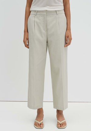 Frau trägt beige Hosen mit weitem Bein und beige Sandalen mit offenen Zehen, steht vor einem einfarbigen hellen Hintergrund.