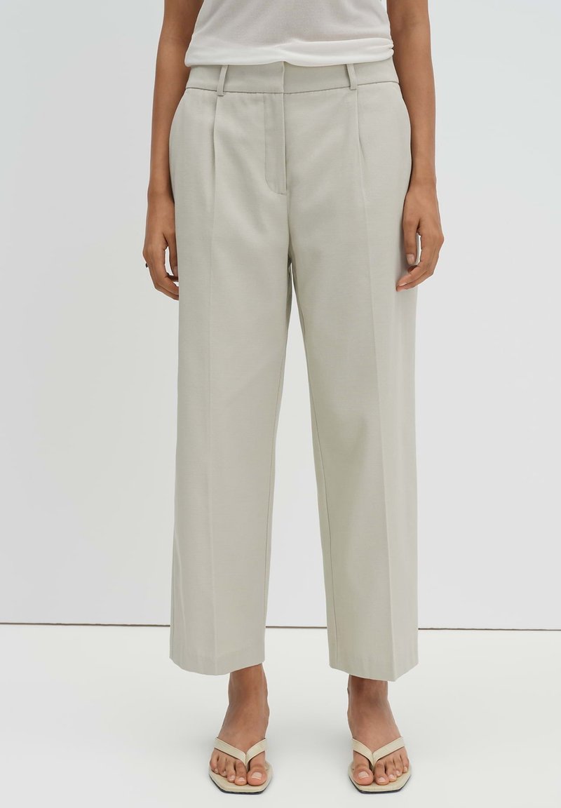 Donna che indossa pantaloni beige a gamba larga e sandali beige aperti davanti, in piedi contro uno sfondo chiaro e semplice.