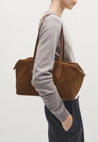 Sac fourre-tout en daim marron avec de longues poignées, de forme rectangulaire et avec une fermeture éclair. Le matériau a une texture douce, exposé aux côtés d'un pull gris.