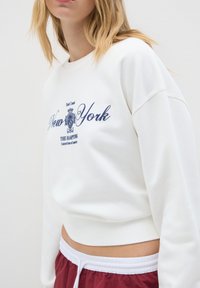 Mujer vestida con un suéter corto blanco con el texto "New York" y pantalones cortos burdeos con cintura elástica, mostrada de cuello a cintura.