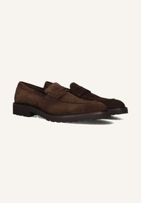 Een paar bruin suède leren loafers met gestikte details en rubberen zolen op een witte achtergrond.