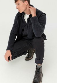 Dunkelgraue Zip-Fleecejacke über einem weißen Shirt, kombiniert mit schwarzen Hosen und schwarzen Schnürstiefeln. Robuste Textur und enger Schnitt.