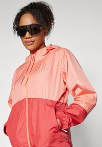 Windbreaker jacket in light pink and coral, featuring a hood, elastic cuffs, and pockets. Made of lightweight, shiny material.