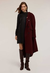 Schwarz gemustertes Kleid mit gefalteten Details, darunter eine burgunderfarbene Jacke. Das Model trägt kniehohe Stiefel aus Schlangenleder in Braun. Goldene Ohrringe setzen Akzente.