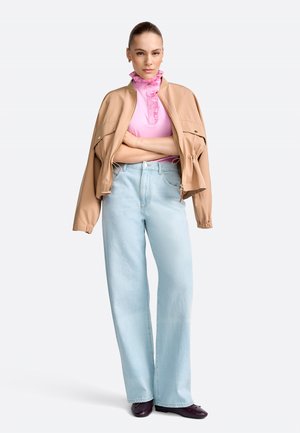 Woman wearing a beige jacket over a pink ruffled top, light blue wide-leg jeans, and dark shoes, standing with arms crossed on white background.