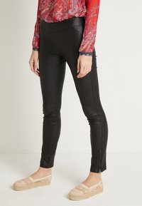 Svarta leggings i konstläder med en stretchig midja, med glidande detaljer vid vristerna. Modellen bär beige espadrillos.