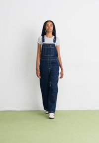 Salopette in denim blu scuro con spalline regolabili, tasche frontali e vestibilità rilassata, abbinata a una maglietta grigia e sneakers bianche.