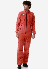 Giacca sportiva arancione con colletto alto, zip frontale e due tasche laterali, abbinata a pantaloni larghi coordinati. Accenti neri sulle tasche.