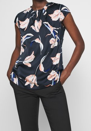 Personne portant un chemisier sans manches noir avec un imprimé floral rose et blanc, associé à un pantalon noir et les mains dans les poches.