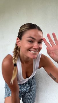 Donna sorridente con capelli intrecciati che saluta, indossando una canotta bianca e pantaloni corti a scacchi, in piedi di fronte a una parete bianca uniforme.