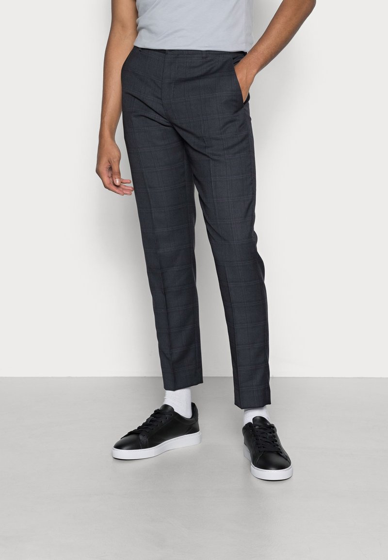 Dunkelkarierte Stoffhosen im Slim-Fit, mit Taschen und einer glatten Textur. Kombiniert mit schwarzen Sneakers und weißen Socken.