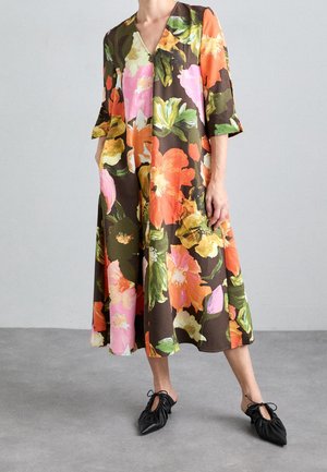Personne portant une robe ample mi-longue avec de grandes impressions florales orange, rose, jaune et verte et des chaussures noires pointues à lacets.