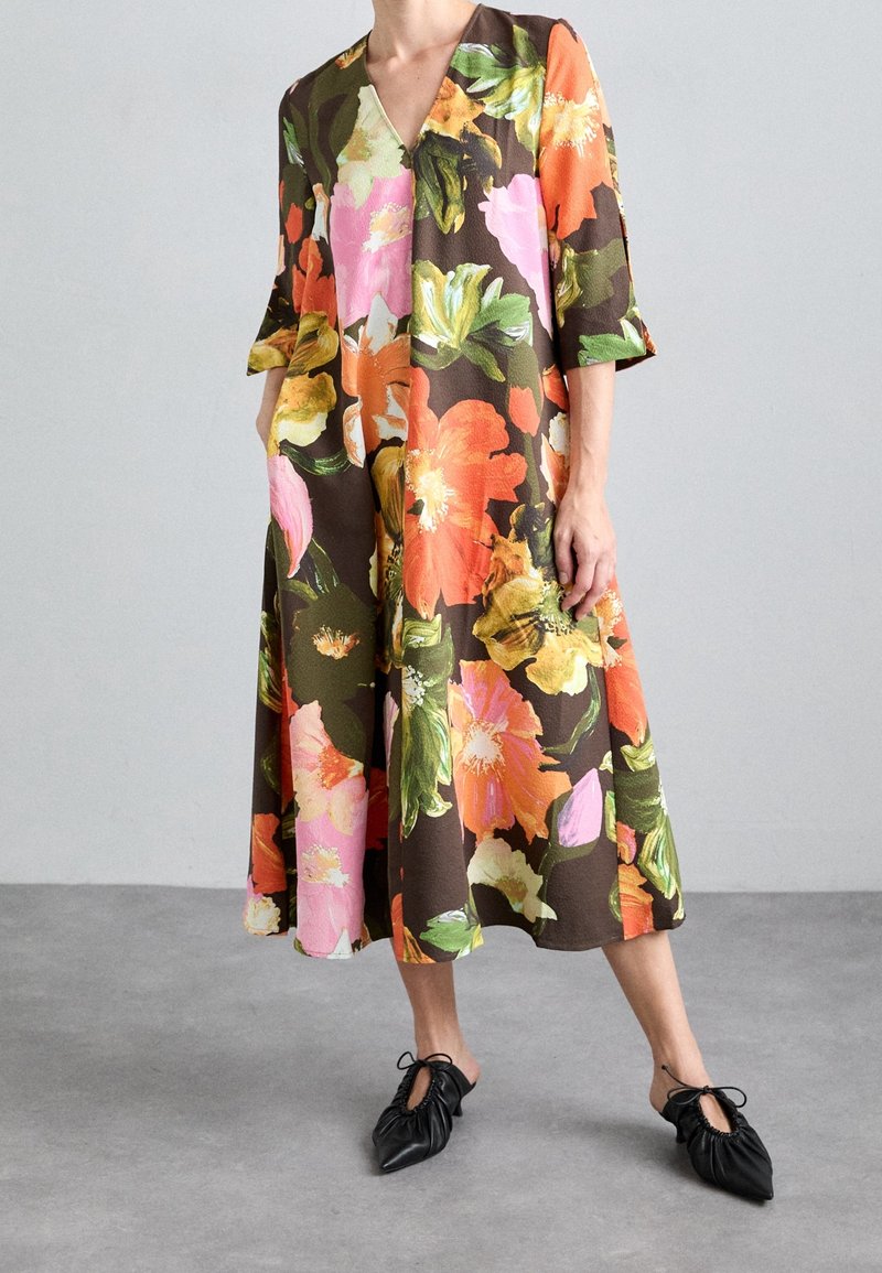 Personne portant une robe ample mi-longue avec de grandes impressions florales orange, rose, jaune et verte et des chaussures noires pointues à lacets.