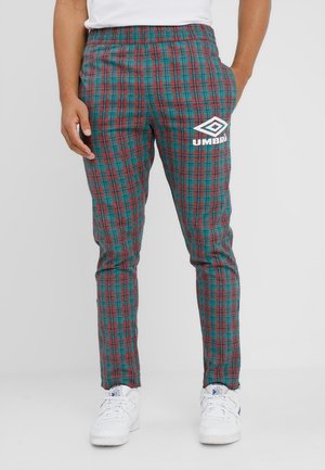 Mand iført tætsiddende rød- og grønternet bukser med Umbro-logo på låret og hvide sneakers med blå detaljer.