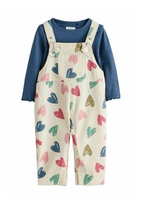 Modra majica z dolgimi rokavi in nazobčanimi manšetami, oblečena pod krem kombinezonom z vzorcem src v roza, zeleni, modri in gorčični barvi.