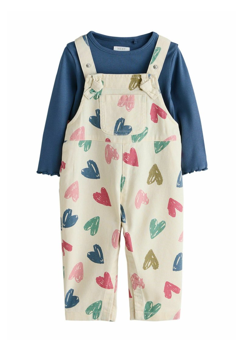 Modra majica z dolgimi rokavi in nazobčanimi manšetami, oblečena pod krem kombinezonom z vzorcem src v roza, zeleni, modri in gorčični barvi.