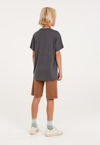 Graues Baumwoll-T-Shirt mit kurzen Ärmeln, kombiniert mit braunen Knie-langen Shorts. Weiße Sneakers und grüne gestreifte Socken vervollständigen das Outfit.