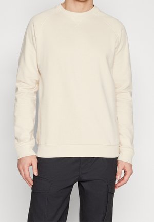 Homme portant un sweat-shirt crème à manches longues et col rond, et un pantalon cargo noir, debout avec les bras relâchés le long du corps.
