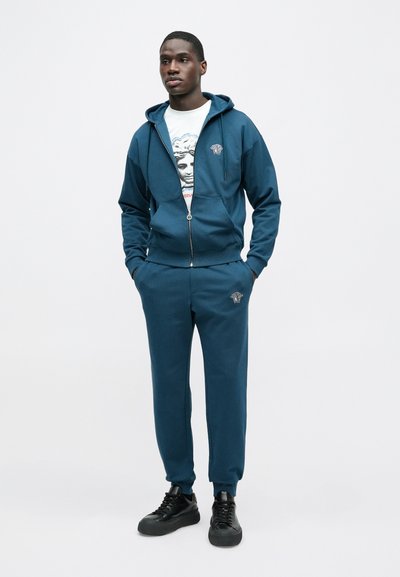 Azúrová mikina na zips s kapucňou, pod ňou sivé tričko s potlačou, zodpovedajúce azúrové joggery a čierne tenisky s plochou podrážkou.