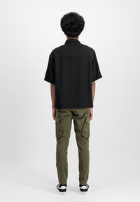 Melnas īsām piedurknēm krekls ar taisnu piegriezi, kombinēts ar olīvzaļām cargo biksēm, kurām ir vairāki kabatas un elastīgas aproces, uzvilktas ar melniem sporta apaviem.