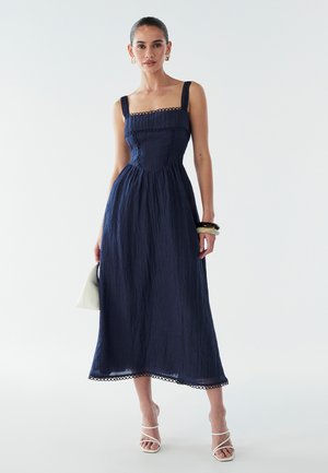 Abito midi blu navy con scollatura quadrata, bordo decorato e vita arricciata. Modella che tiene una borsa bianca e indossa sandali con tacco bianchi.