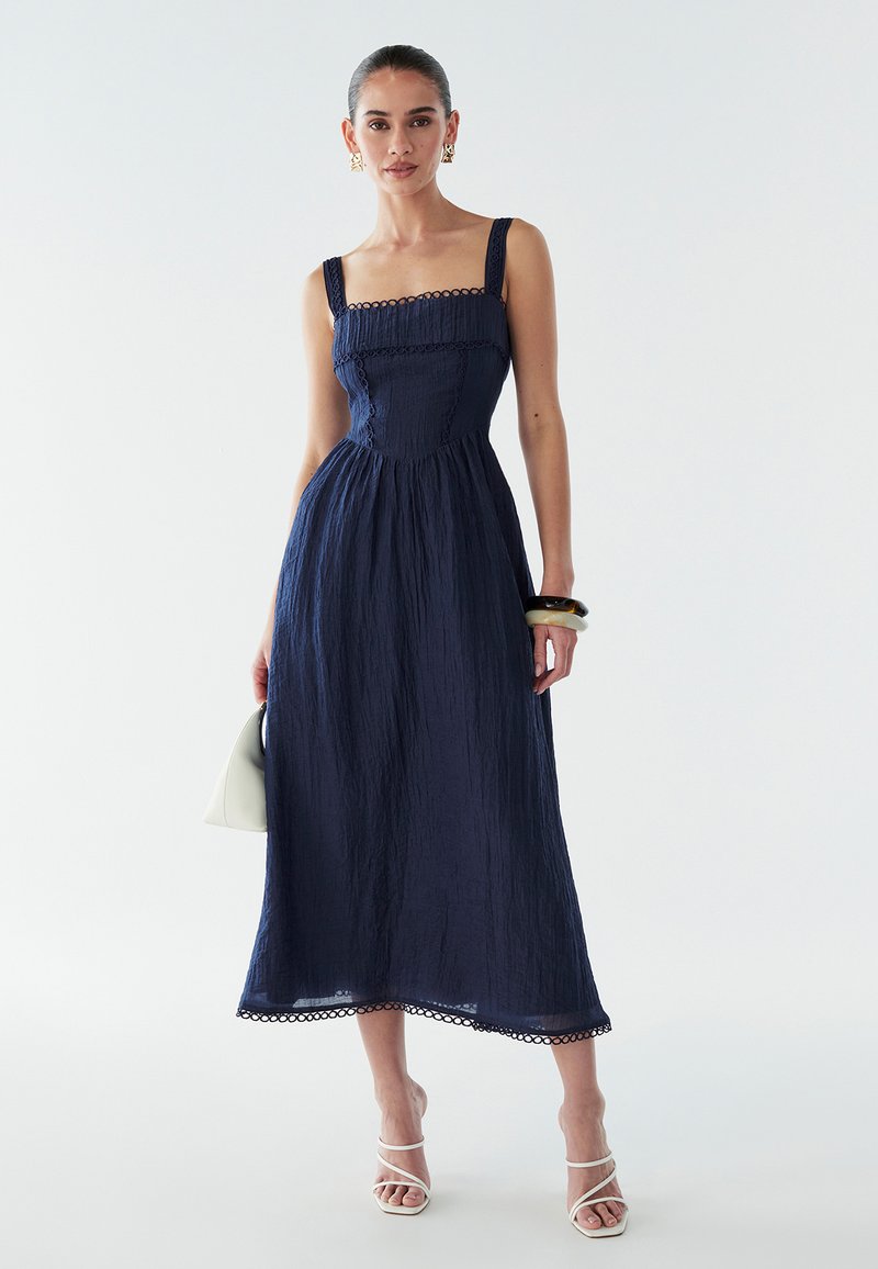 Vestido azul marino de longitud media con tela texturizada, escote cuadrado, ribete de encaje y tirantes delgados. Combinado con un bolso claro y sandalias blancas.