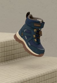 Botte de randonnée montante bleue et marron avec semelle à épaisseur importante, sangle Velcro et lacets élastiques suspendue au-dessus d'escaliers en carrelage beige.