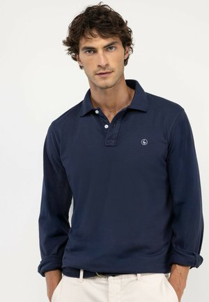 Hombre con cabello rizado y oscuro que lleva una camisa polo azul marino de manga larga y pantalones blancos con las manos en los bolsillos sobre un fondo liso.