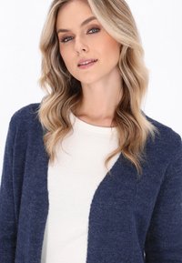 Granatowy sweter z dzianiny z otwartym przodem, prezentujący teksturowane wykończenie. Nosi się go na jasnym, okrągłym dekolcie.