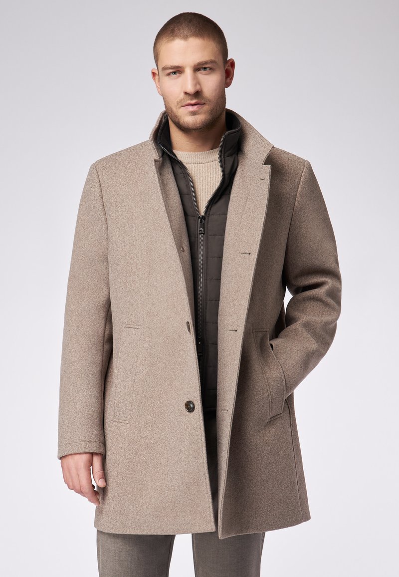 Hellbrauner Wollmischmantel mit Reverskragen, Ein-Knopf-Verschluss, zwei Fronttaschen, getragen über einer schwarzen Steppjacke.