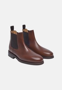 Bottines Chelsea en cuir marron avec des panneaux élastiques latéraux noirs, à bout arrondi et à talon bas empilé. Texture lisse avec des coutures minimales.