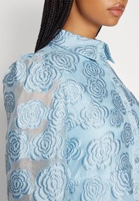 Kvinna med flätat hår iförd en genomskinlig blå blus broderad med blommönster och knäppt framtill, sedd från sidan.