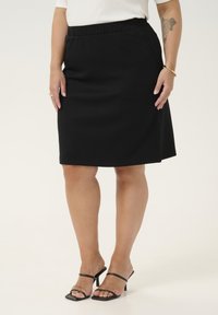 Jupe noire longueur genou avec taille élastique et poches latérales, fabriquée en tissu doux. Portée avec des talons noirs à lanières. Texture lisse.
