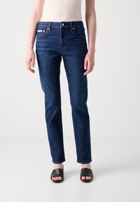 Jeans de ganga azul escura com um design de perna reta, apresentando bolsos frontais e fecho de botão. Usados com sandálias de slide pretas.