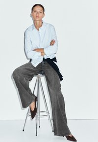 Frau sitzt auf einem Metallhocker, trägt ein hellblaues, geknöpftes Hemd, weite graue Hose, dunkle Schuhe, Arme verschränkt, vor einem einfarbigen hellen Hintergrund.