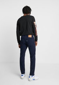 Svart långärmad tröja med färgglad grafik på ärmen, matchad med marinblå manchesterbyxor och vita sneakers med blå detaljer.