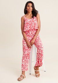 Combinaison rouge à motifs avec un haut superposé sans manches et des jambes fuselées. Présente de grands motifs de feuilles blanches, assortie à des sandales beiges.