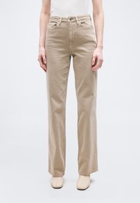 Άτομο που φοράει μπεζ τζιν παντελόνι με ίσια γραμμή και μπεζ μπότες αστραγάλου με τετράγωνη μύτη, σκυθρωπό σε απλό φωτεινό φόντο.