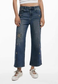 Sinised laia lõikega teksad tikanditega, millel on kõrge vöökoht ja nööpkinnitus. Kangas on pehme tekstuuriga ja veidi tuhmunud.