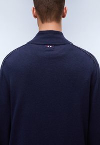 Vue de dos d'une personne portant un pull en maille bleu marine avec un col montant et une petite étiquette rouge, blanche et bleue au niveau du cou.