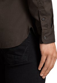 Camisa de manga larga de color marrón oscuro con puños abotonados, tela texturizada, dobladillo redondeado y silueta ajustada, combinada con vaqueros negros que presentan un detalle de bolsillo.