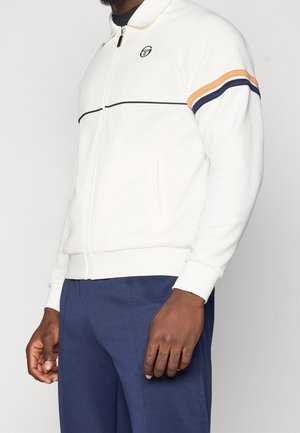 Personne portant une veste blanche zippée avec des rayures orange et bleu marine sur la manche et un pantalon bleu marine, visible des épaules jusqu’au milieu de la cuisse.