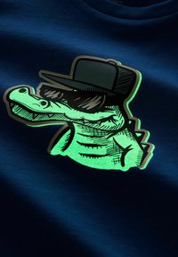 Navy zils T-krekls ar spīdošu zaļu krokodila grafiku, kuram ir saulesbrilles un cepure, ar detalizētu līniju mākslu un teksturētu audumu.