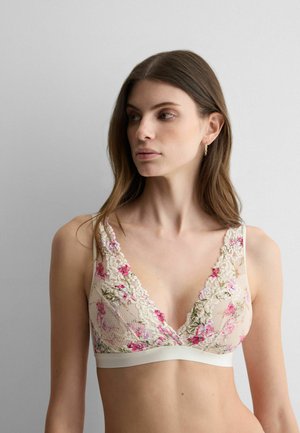 Junge Frau mit langen braunen Haaren, die ein florales Spitzen-Bralette trägt, blickt nach links vor einem schlichten grauen Hintergrund.