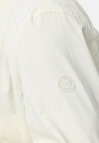 Let lys jakke i hvid farve med en glat tekstur, en syet skuldersøm og et rundt hvidt logo-patch på overarmen.