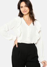 White blouse with ruffled details along the shoulders, textured fabric, V-neckline, and button closure, paired with black high-waisted pants.