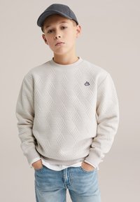 Világosszürke pulóver texturált hatszögmintával, bordázott nyakkivágással és mandzsettákkal, fehér ing fölött viselve. Sötétszürke sapka egészíti ki a megjelenést.