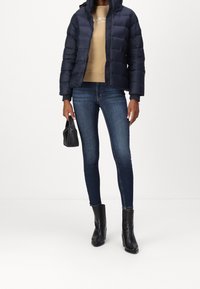 Giacca a vento blu navy, felpa beige, jeans skinny blu scuro e stivaletti neri con tacco. Il modello tiene una piccola borsa nera.