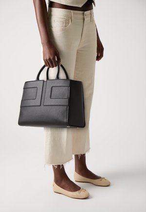 Personne tenant un grand sac à main en cuir noir texturé, portant un jean crème court avec des ourlets effilochés et des ballerines beiges avec de petits nœuds.
